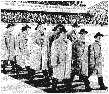 Die Herren der österreichischen Olympiamannschaft 1956, bekleidet mit dunklen Hüten, langen hellen Mänteln und dunkeln Hosen beim Einzug ins Stadion.